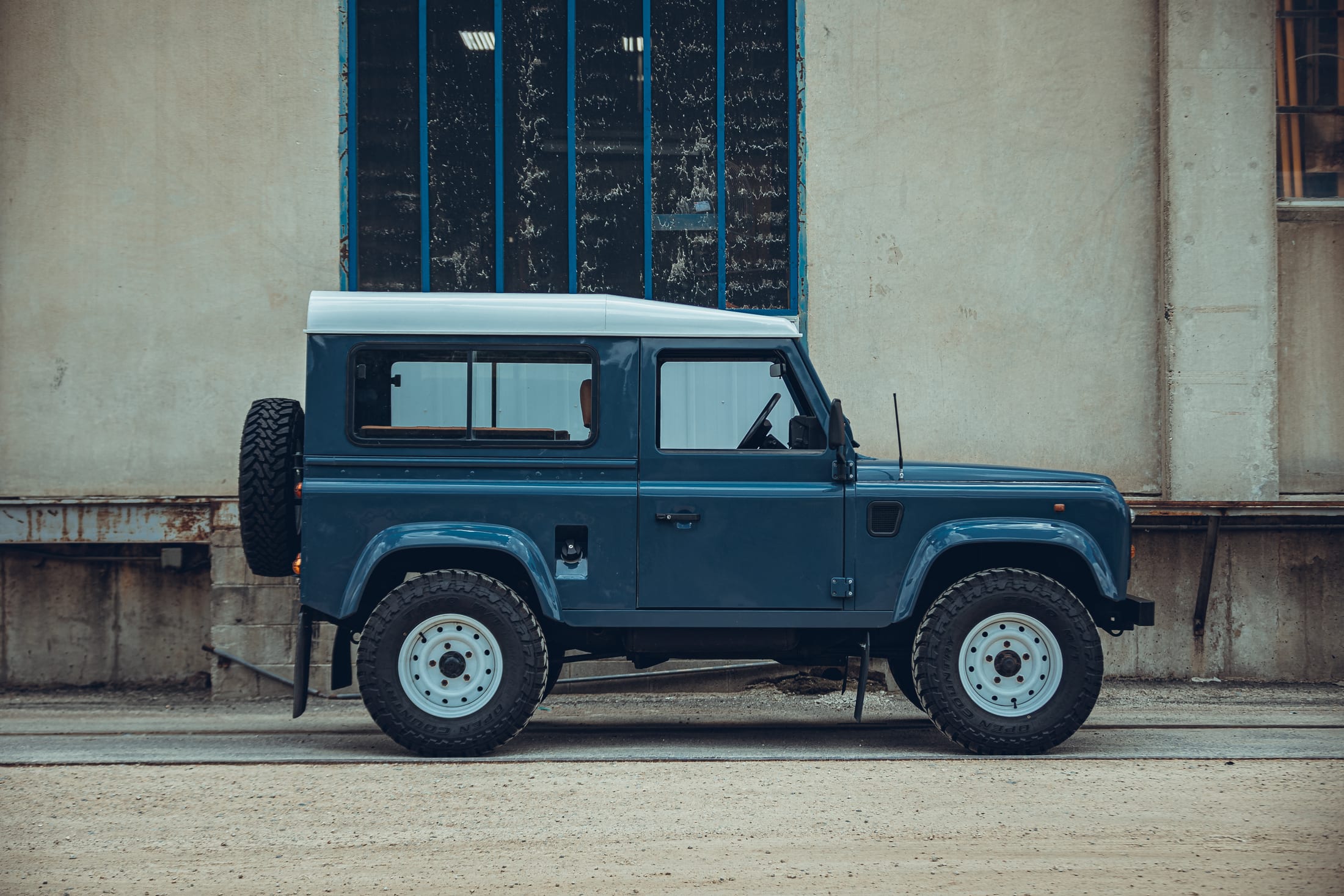 A blue Land Rover Defender 90 parked against a weathered workshop wall